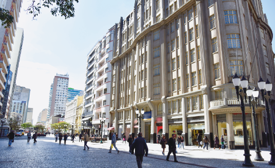 Imagem: Fotografia. Destaque de uma via onde há muitas pessoas caminhando. Ao fundo, um edifício alto e amplo com muitas janelas. Do lado direito, uma fileira de postes de iluminação. Fim da imagem.