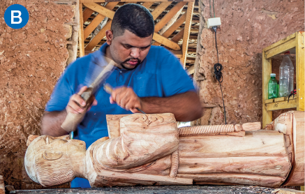 Imagem: B. Fotografia. Em um local com paredes de barro, um homem de camiseta está de pé ao lado de uma superfície na qual há uma escultura de madeira. Ele manipula dois instrumentos em suas mãos. Fim da imagem.