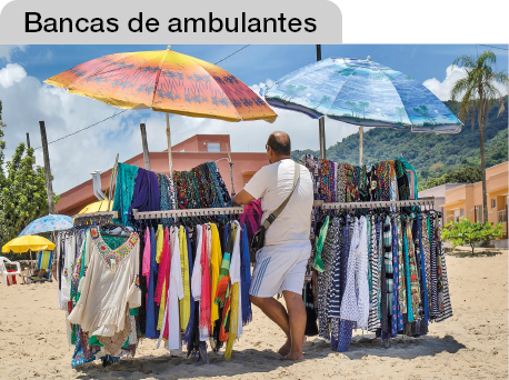 Imagem: Fotografia. Na areia da praia, um homem está de costas e de pé encostado em uma arara com muitas roupas de banho e dois guarda-sóis abertos. Ao fundo, casas e a serra. Fim da imagem.