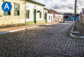 Imagem: A. Fotografia. Destaque de uma rua com paralelepípedos e calçadas estreitas com casas térreas com portas e janelas voltadas para via.  Fim da imagem.