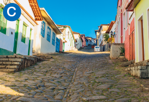 Imagem: C. Fotografia. Destaque de uma ladeira estreita feita com pedras. As calçadas são bem estreitas, as casas são térreas e com portas e janelas voltadas diretamente para a rua.  Fim da imagem.