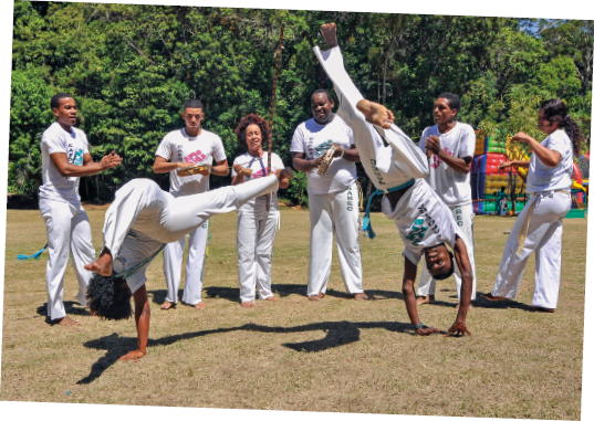 Imagem: Fotografia. Uma roda de capoeira em área gramada composta por oito pessoas. Ao centro, duas pessoas jogam, com as mãos no chão e os pés para o alto, e ao redor, o restante bate palmas e toca instrumentos. Eles estão descalços, usam camiseta e calça branca. Ao fundo, muitas árvores.  Fim da imagem.