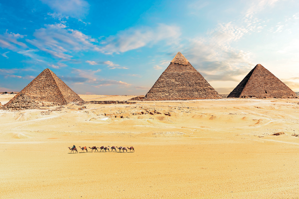Imagem: Fotografia. Plano aberto de uma extensa faixa de areia. Em primeiro plano, um homem a camelo está diante de uma fileira com outros seis animais. Ao fundo, três robustas construções piramidais. No horizonte, o céu azul e nuvens.  Fim da imagem.