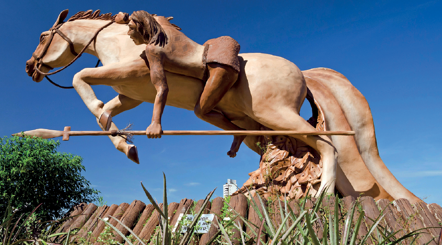 Imagem: Fotografia. Destaque de uma escultura de um homem seminu segurando uma lança e com o corpo voltado para frente enquanto cavalga sobre um cavalo.  Fim da imagem.