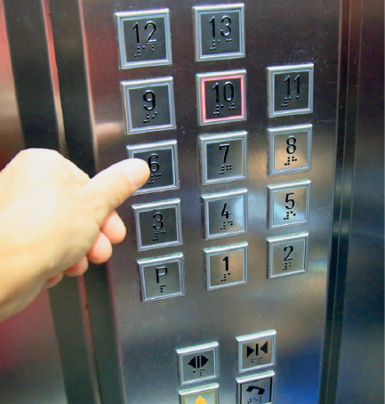 Imagem: Fotografia. Destaque da mão de uma pessoa que manipula botões de um elevador. Fim da imagem.
