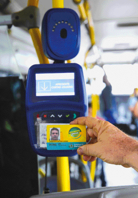 Imagem: Fotografia. No interior de um ônibus, destaque da mão de uma pessoa que aproxima um cartão de plástico de um aparelho eletrônico. Fim da imagem.