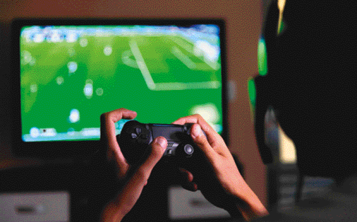 Imagem: Fotografia. Em primeiro plano, destaque da mão de uma pessoa que manipula um controle remoto de videogame. Ao fundo, um monitor amplo com a imagem de um estádio de futebol. Fim da imagem.
