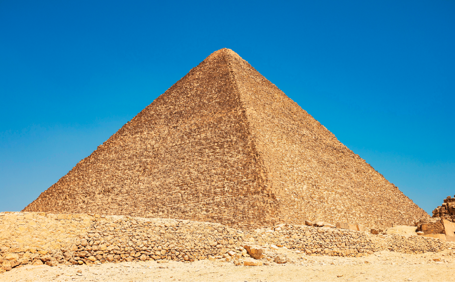 Imagem: Fotografia. Destaque de uma robusta construção piramidal em meio à área desértica. No horizonte, céu azul. Fim da imagem.