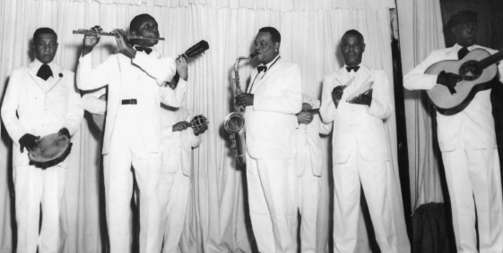 Imagem: Fotografia em preto e branco. Um grupo composto por seis homens negros de terno claro tocam instrumentos de pé. Entre os instrumentos estão pandeiro, flauta transversal, violão, saxofone e prato.  Fim da imagem.