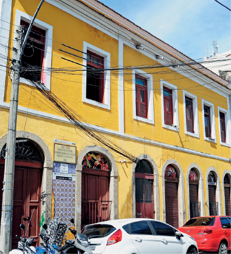 Imagem: Fotografia. Destaque de uma fachada assobradada amarela com muitas janelas e portas arqueadas em tom avermelhado. Na rua, há carros e motos estacionados e poste de iluminação. Fim da imagem.