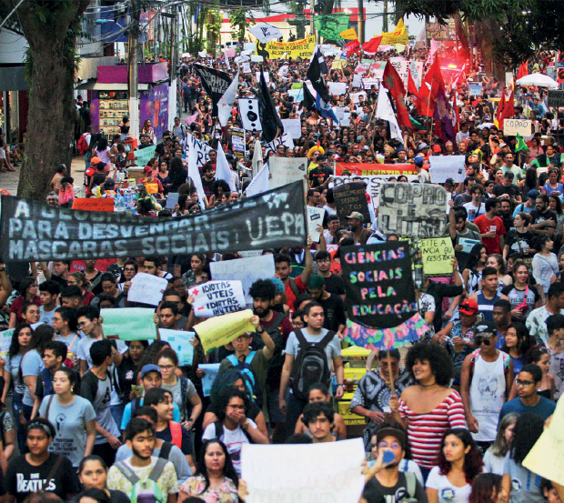 Imagem: Fotografia. Destaque de uma multidão que caminha em uma rua segurando cartazes e faixas. Dentre os dizeres, estão “CIÊNCIAS SOCIAIS PELA EDUCAÇÃO”. Fim da imagem.