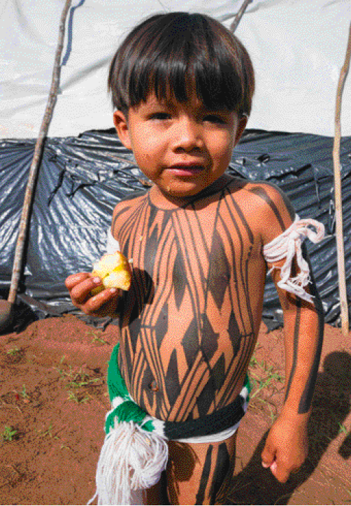 Imagem: Fotografia. Um menino indígena está de pé sem camisa e come uma maçã. Ele tem cabelo liso preto e comprido, olhos ligeiramente puxados, pele parda, usa adereços com linhas nos braços e na cintura e tem e tem o corpo pintado com tinta preta. Fim da imagem.