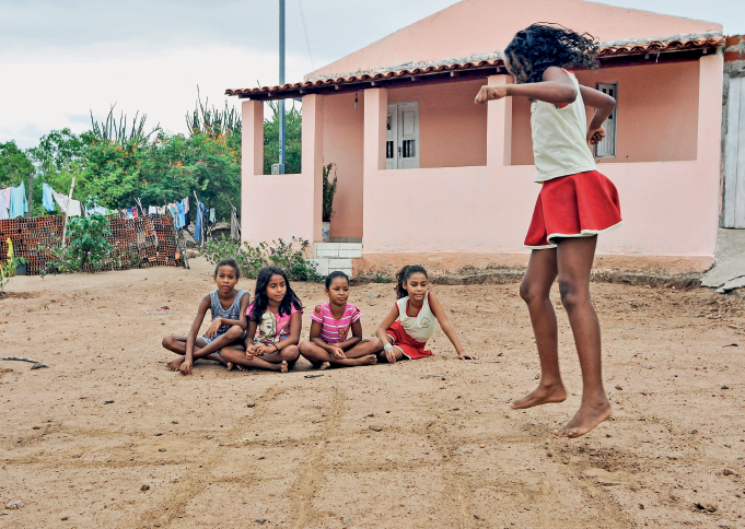 Imagem: Fotografia. Em um local com solo arenoso diante de uma pequena casa, cinco crianças brincam.  Quatro estão sentadas lado a lado e uma delas pula uma amarelinha contornada no chão.  Fim da imagem.