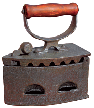 Imagem: Fotografia. Um ferro de passar que apresenta corpo robusto com abertura superior com rebarbas e pegador de madeira.  Fim da imagem.