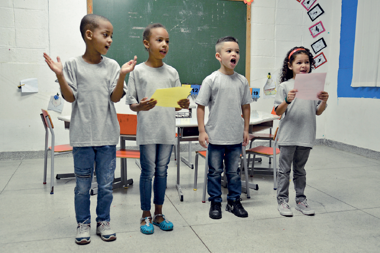 Imagem: Fotografia. No interior de uma sala de aula, quatro crianças uniformizadas estão de pé diante de uma mesa próximo à lousa e se apresentam. Estão enfileirados lado a lado, os meninos falam e um deles bate palma e as meninas falam segurando folhas de papel. Fim da imagem.