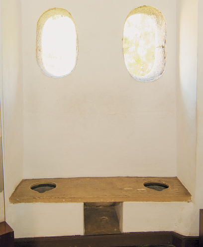 Imagem: Fotografia. Interior de um espaço que apresenta um assento de alvenaria com duas aberturas ovalares na superfície. Na parede, janela com duas aberturas ovalares vazadas.  Fim da imagem.