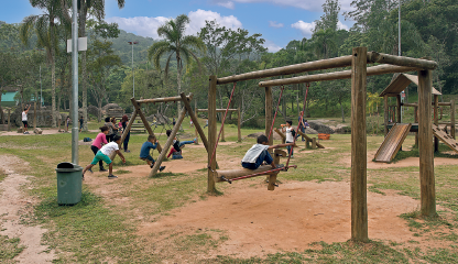 Imagem: Fotografia. Área aberta com chão de terra e grama onde brincam crianças em um parquinho com estrutura de madeira. Ao fundo, vegetação. Fim da imagem.