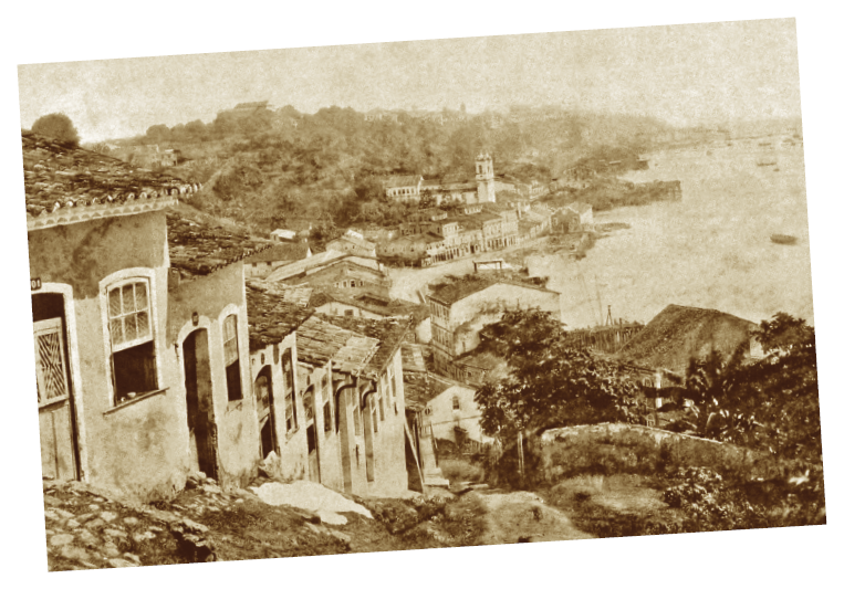 Imagem: Fotografia sépia. Destaque de uma ladeira com chão de terra, arbustos e árvores de um lado e casebres com portas e janelas voltadas diretamente para rua do outro. Ao fundo, construções à beira-mar, com algumas embarcações, e vegetação. Fim da imagem.