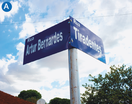 Imagem: A. Fotografia. Destaque de um poste com placas do encontro de duas ruas: Artur Bernardes e Tiradentes. Fim da imagem.