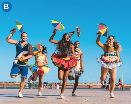 Imagem: B. Fotografia. Em um espaço aberto, um grupo de cinco pessoas, homens e mulheres, se apresenta. Todos seguram pequenas sombrinhas coloridas e usam tênis. Os rapazes usam camiseta regata e shorts e as meninas usam regata e saia curta rodada e volumosa.  Fim da imagem.