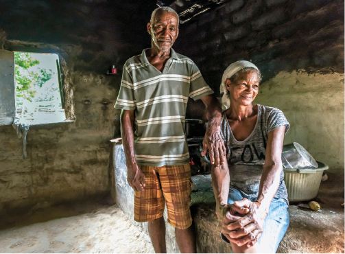 Imagem: Fotografia. Um senhor e uma senhora negros posam no interior de um casebre. Ela tem um lenço na cabeça, usa camiseta e shorts e olha para o lado e está sentada com as duas mãos no joelho. Ele está de pé ao seu lado, usa camiseta e shorts, apoia o braço no seu ombro direito e olha para a câmera.  Fim da imagem.