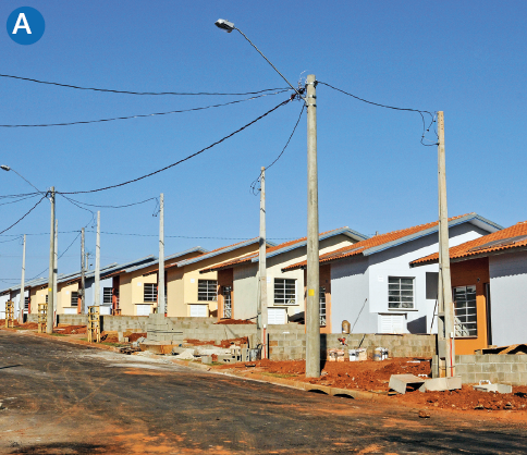 Imagem: A. Fotografia. Destaque de uma rua com pavimento recente, postes de iluminação e pequenas casas semelhantes em construção lado a lado. Há terra e blocos nas calçadas.  Fim da imagem.