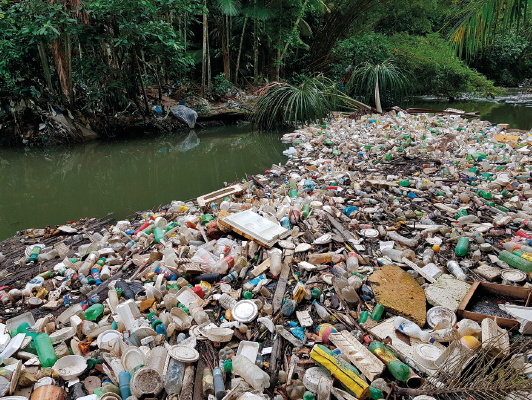 Imagem: Fotografia. Destaque de um amontoado de lixo, sendo a maioria material plástico, à margem de um rio. Do outro lado da margem, há vegetação.  Fim da imagem.