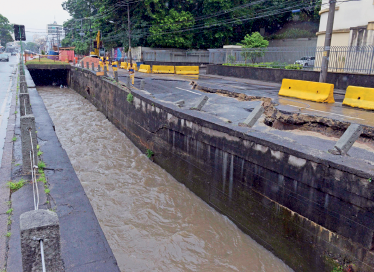 Imagem: Fotografia. Destaque de um rio que corre em um canal estreito e urbano.  Fim da imagem.