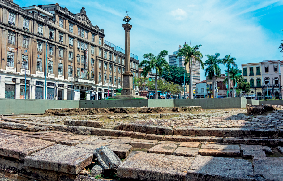 Imagem: Fotografia. Plano aberto de uma área urbana com destaque para o chão com grandes pedras e uma área descoberta. No local, há um obelisco e ao fundo estão alguns prédios.  Fim da imagem.