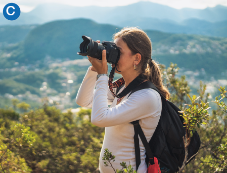 Imagem: C. Uma mulher de camiseta de frio e mochila nas costas fotografa com uma câmera profissional. Ao fundo, morro e cidade.  Fim da imagem.