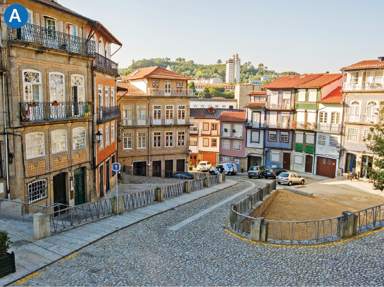 Imagem: A. Fotografia. Plano aberto do centro de uma cidade que apresenta uma rampa com pequenas pedras em mosaicos e ruas que apresentam construções assobradadas com amplas portas com sacadas, parapeito com arabescos e portas com acesso diretamente à rua. Na via, há alguns carros estacionados.  Fim da imagem.