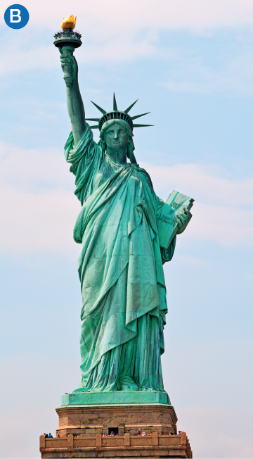 Imagem: B. Fotografia. Uma estátua grande representada por uma mulher de pé que usa uma roupa comprida e larga, segura um livro abraçado na mão esquerda e segura uma tocha na mão direita com o braço estendido. Na cabeça, usa uma coroa com sete pontas.  Fim da imagem.