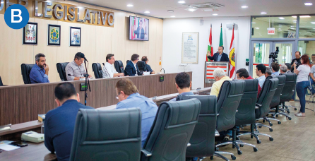 Imagem: B. Fotografia. Destaque do interior de uma sala onde há pessoas sentadas enfileiradas lado a lado diante de uma comprida bancada. À frente deles, homens estão sentados diante de microfones. No canto direito, está um outro homem à frente de três mastros com bandeiras. Fim da imagem.