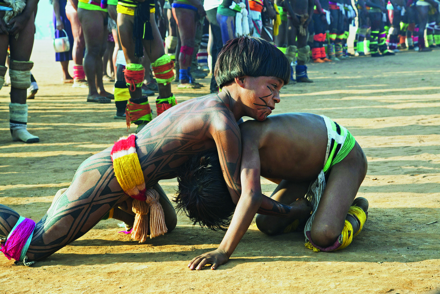 Fotografia. Dois meninos indígenas ajoelhados, com o corpo e o rosto pintados. O menino da esquerda está com as mãos para baixo e a cabeça sobre as costas do outro menino.