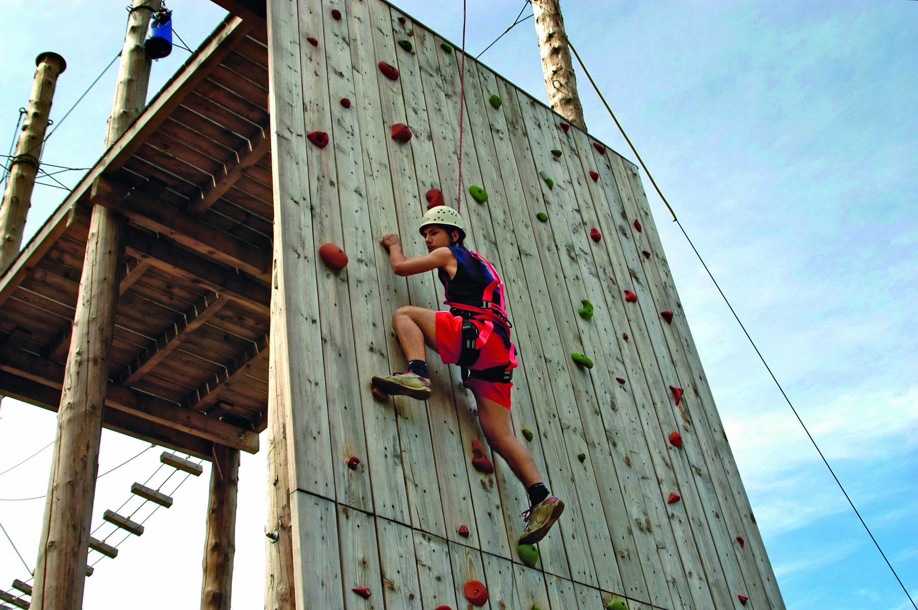 Fotografia. Rapaz de camiseta, bermuda e capacete escalando uma parede. Ele está preso a uma corda e se agarra a pontos de apoio fixados na parede.