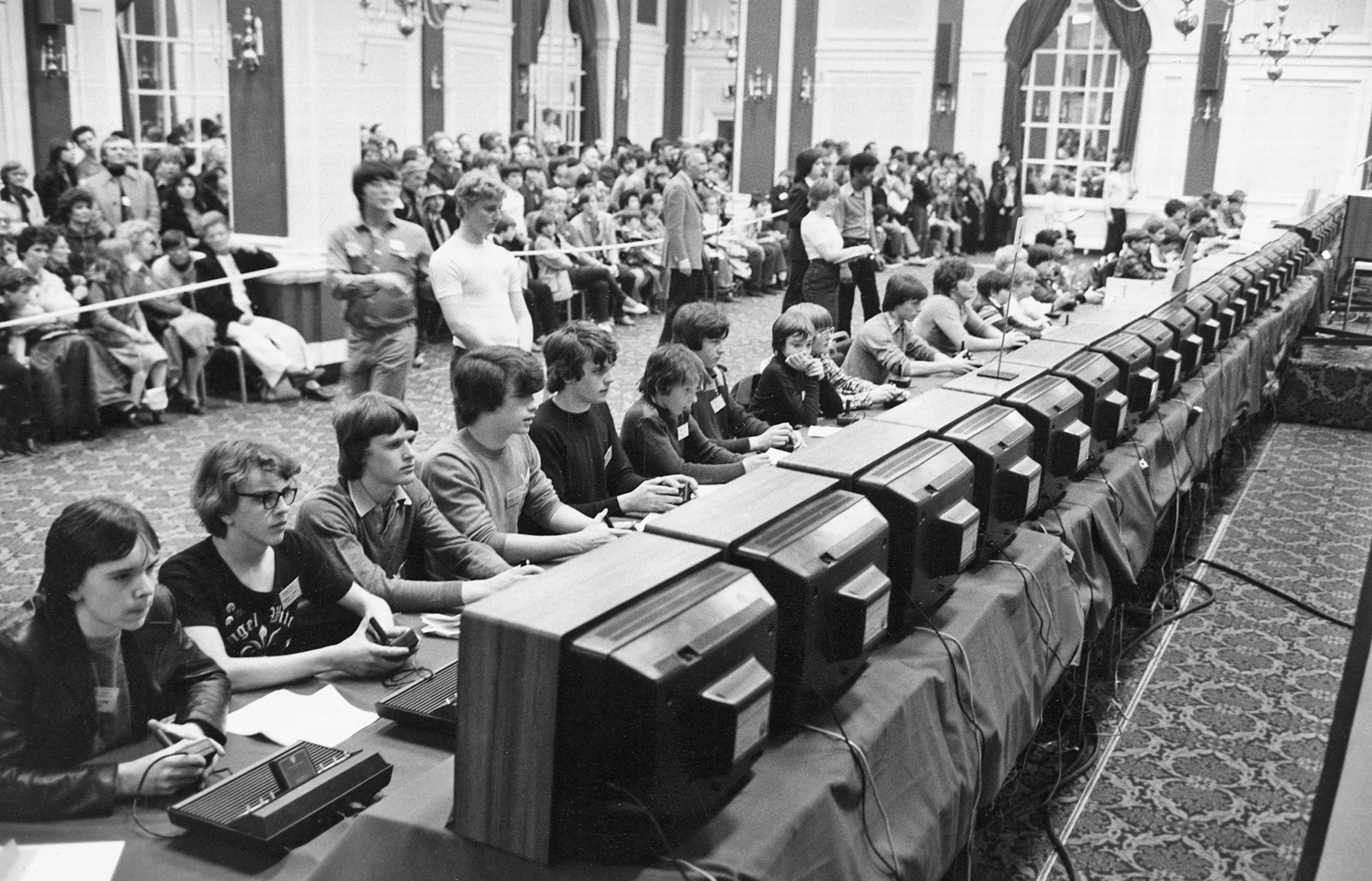 Fotografia em preto e branco. Vários jovens, sentados um ao lado do outro, de frente para um computador. Atrás, uma faixa os separa das pessoas que estão acompanhando o campeonato de videogame.