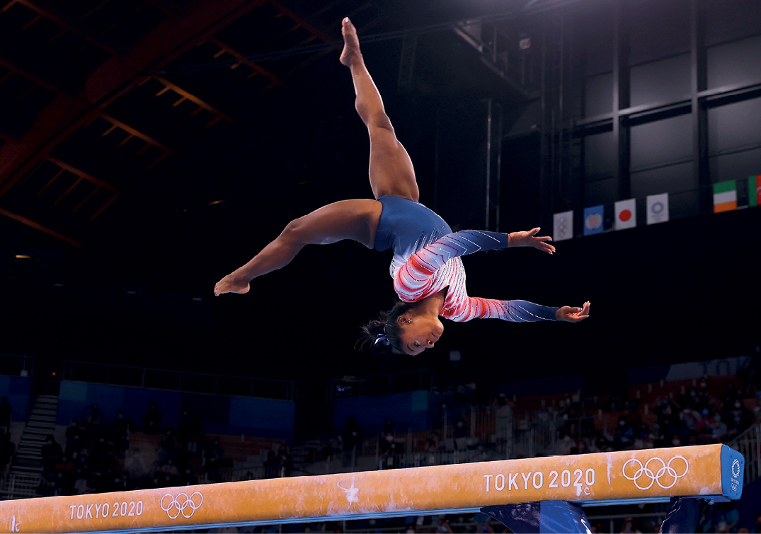 Fotografia. De uniforme azul, branco e vermelho, a ginasta negra Simone Biles está saltando sobre uma trave. As pernas estão voltadas para cima, os braços estendidos para a frente e a cabeça para baixo.