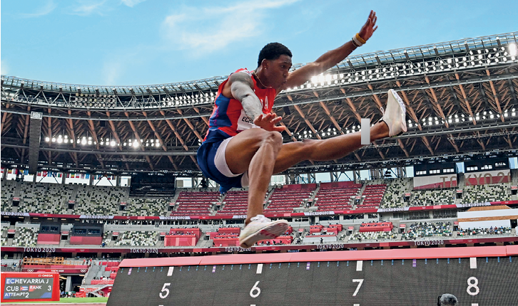 Fotografia. De uniforme vermelho e azul, o atleta negro Juan Miguel salta com o joelho direito flexionado e a perna esquerda estendida, o braço esquerdo para cima e o braço direito para a frente.