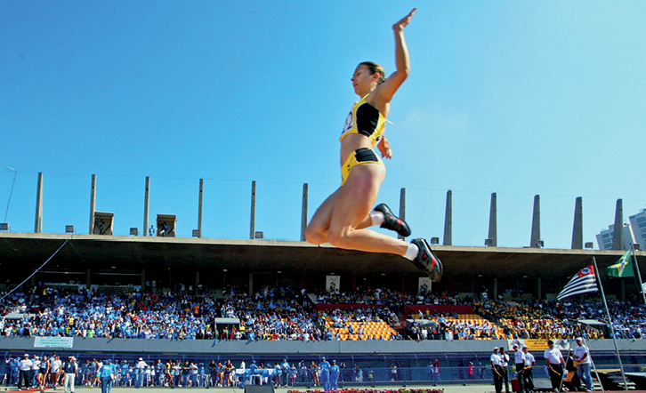 Fotografia. De uniforme amarelo, a atleta branca Maurren Maggi salta com os joelhos flexionados e as pernas para trás.