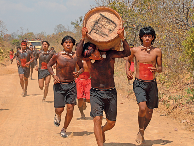 Fotografia.  Um grupo de jovens indígenas com o corpo pintado, uns de preto, outros de vermelho, corre em uma rua de terra. O indígena que está à frente carrega uma tora de madeira nas costas.