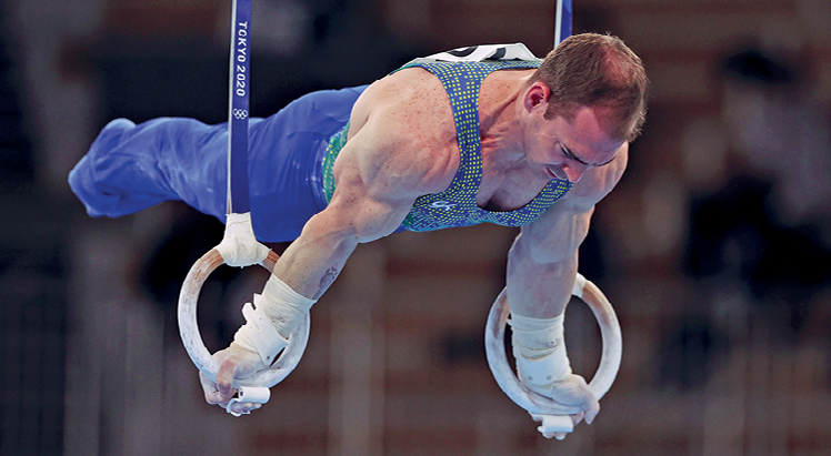 Fotografia. O ginasta branco Arthur Zanetti, de collant azul, segura em duas argolas suspensas, mantendo o corpo estático, na horizontal, usando para isso a força dos braços.