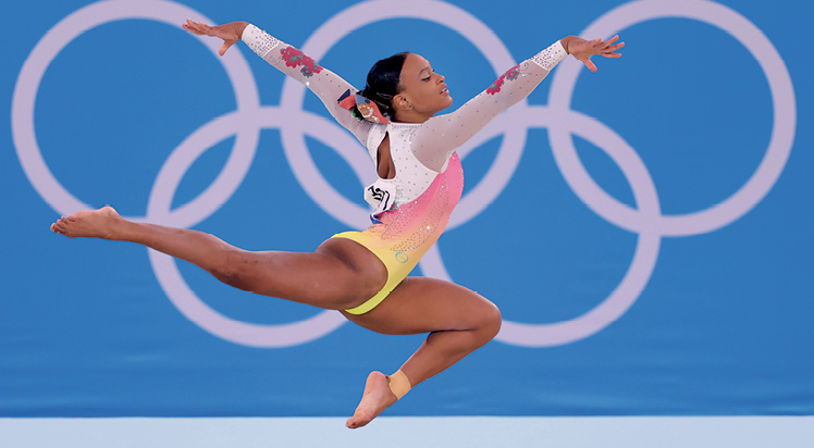 Fotografia. A ginasta negra Rebeca Andrade, de collant amarelo, rosa e branco, salta de perfil com o braço direito para a frente, o braço esquerdo para trás, perna direita estendida para trás e perna esquerda com o joelho flexionado próximo ao corpo.