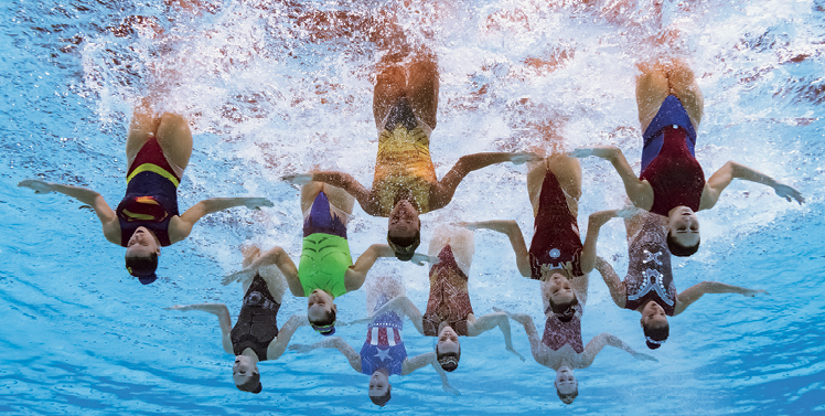 Fotografia subaquática. Grupo de dez moças em uma piscina, de cabeça para baixo e braços estendidos na horizontal.
Somente as pernas ficam de fora e todas fazem o mesmo movimento. Elas usam maiôs coloridos.