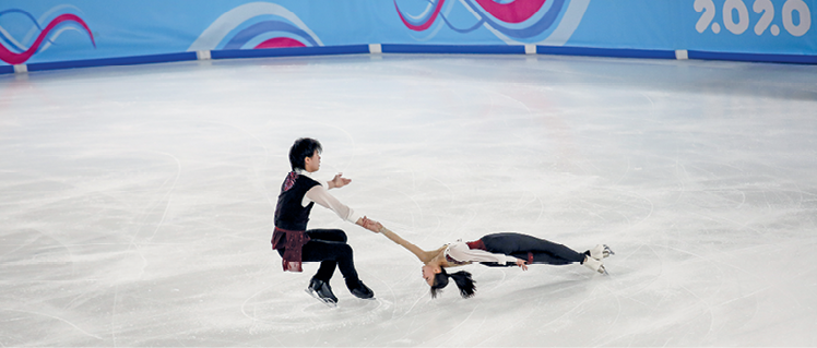 Fotografia. Casal chinês com roupa preta e patins se apresenta sobre o gelo. O rapaz está agachado e segura a mão da moça, que está com o corpo estendido, quase tocando o gelo.
