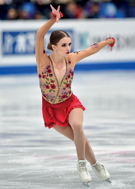 Fotografia. De collant florido e saia vermelha, a ginasta branca Isadora Willliams se apresenta sobre patins no gelo. Ela está com os braços para cima e a perna esquerda cruzada na frente da perna direita.