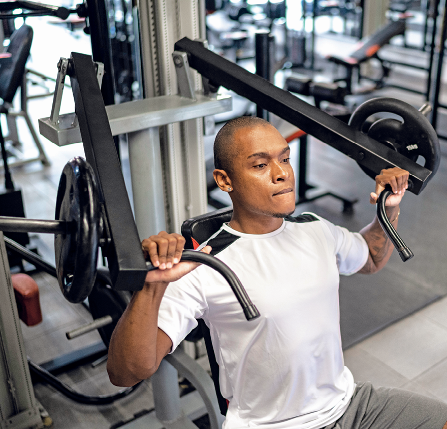 Fotografia. Homem de cabelo raspado, camiseta branca e bermuda cinza. Ele está se exercitando em uma academia, segurando com as mãos uma barra com pesos.