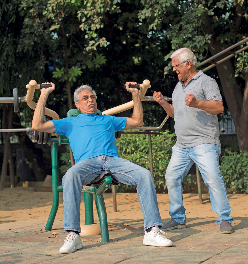 Fotografia. Homem de meia-idade, de cabelo grisalho, em uma praça exercitando-se em aparelho de ferro composto por acento e levantamento de braço. À direita, outro homem, também de meia-idade, de cabelo grisalho, incentiva o amigo.