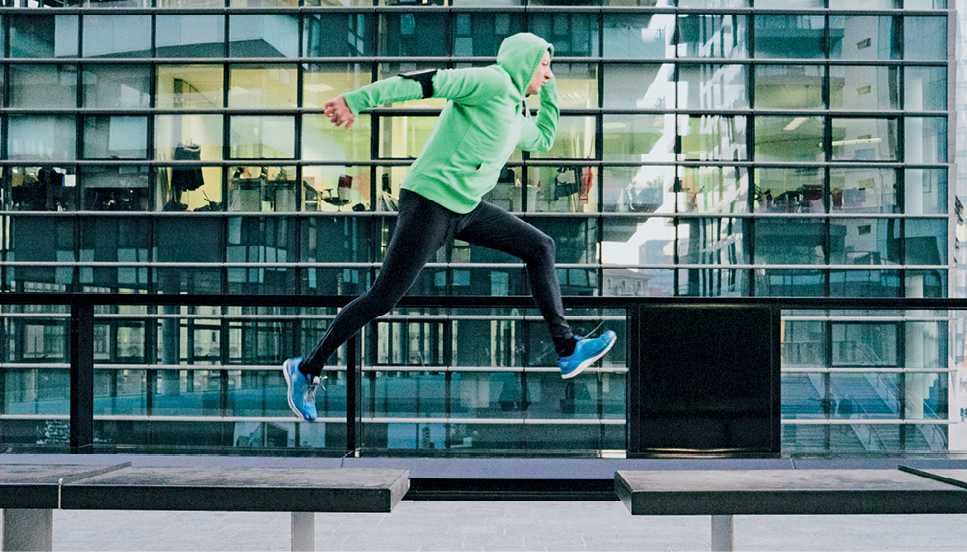 Fotografia. Homem com agasalho verde com capuz, calça preta e tênis. Ele está saltando entre o vão de dois bancos. Atrás, prédio com parede de vidro.