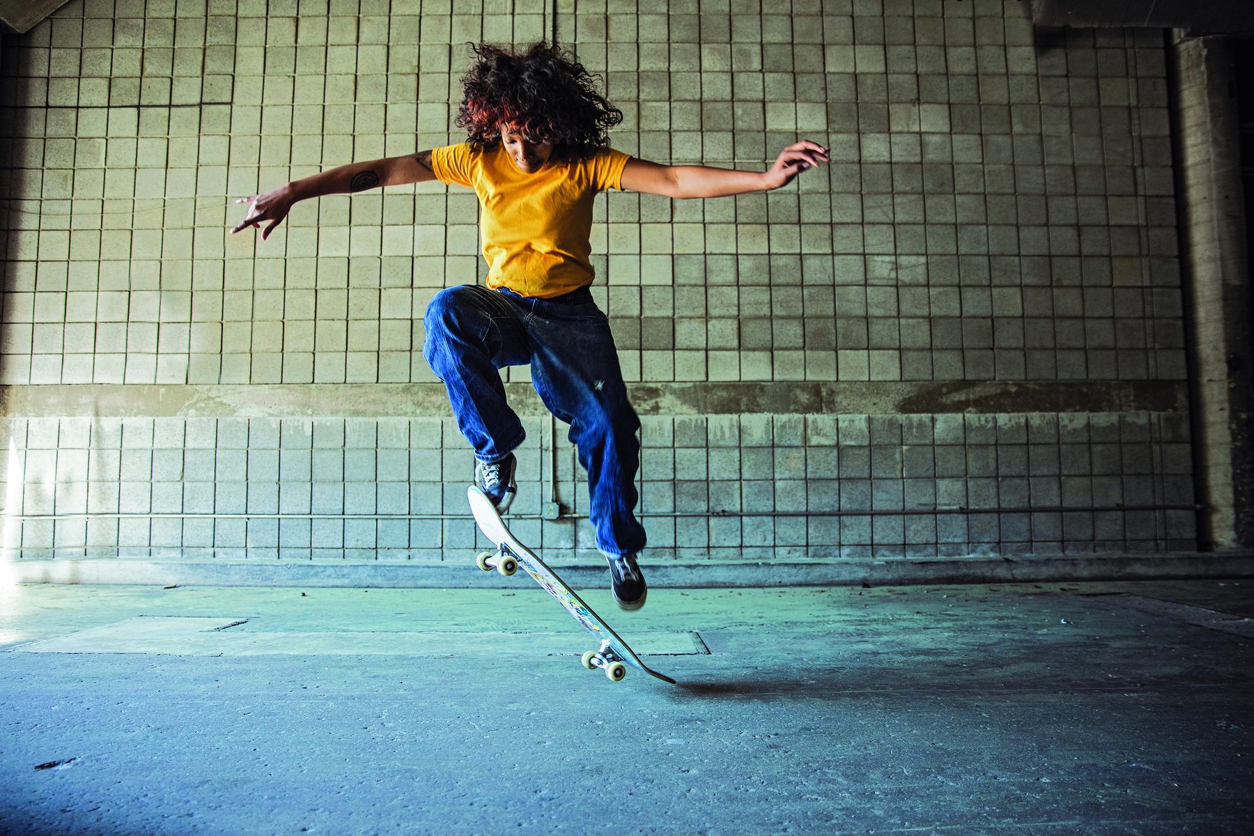 Fotografia. Mulher usando camiseta amarela, calça jeans e tênis. Ela está saltando sobre um skate com a perna direita dobrada e os braços abertos.