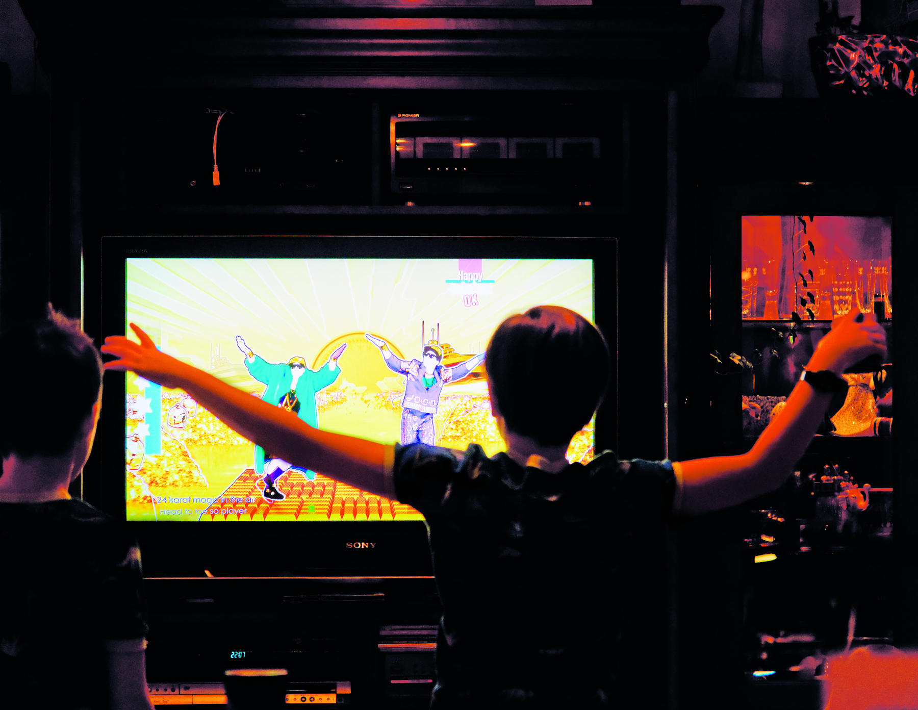 Fotografia. Uma criança está em pé e com os braços abertos de frente para uma televisão; na tela há um jogo com duas pessoas dançando.